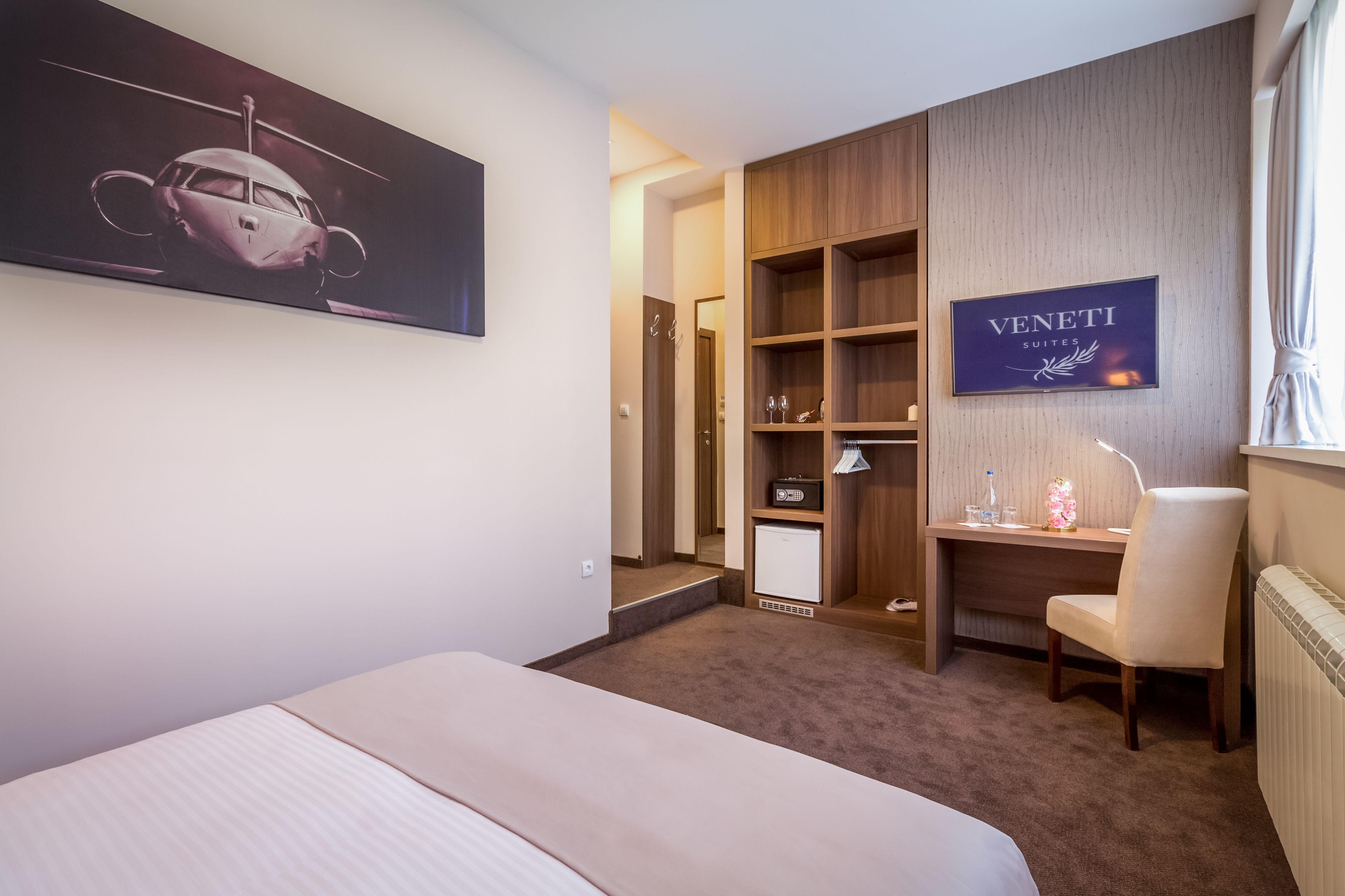 A hotel room with a bed, a desk, a chair, a wardrobe, and a painting on the wall.