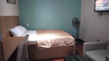 A room with a bed, a desk, a chair, a television, and a standing fan.