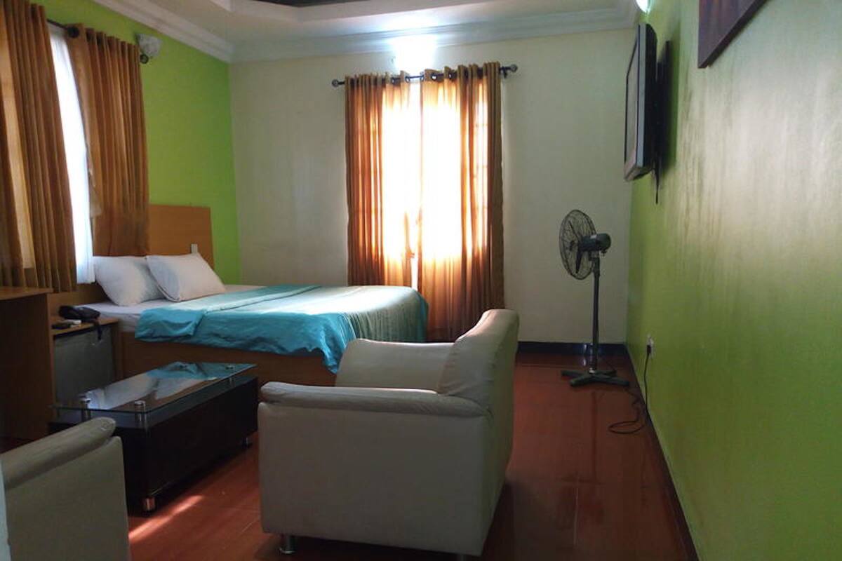 Ein Hotelzimmer mit Bett, Sessel, Schreibtisch, Ventilator und einem Fenster mit Vorhängen.