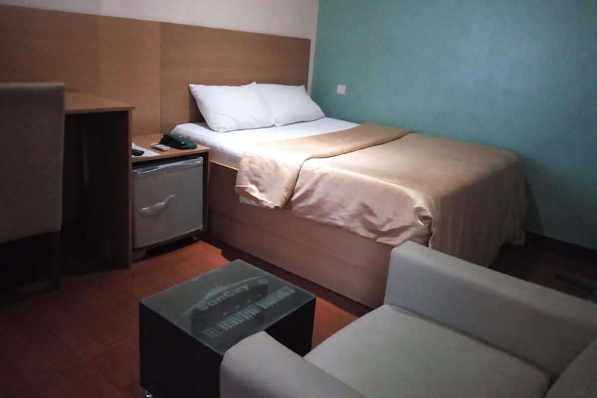Ein kleines, ordentlich eingerichtetes Hotelzimmer mit einem Einzelbett, einem Nachttisch mit Lampe, einem Sessel und einem kleinen Tisch mit Tastatur.