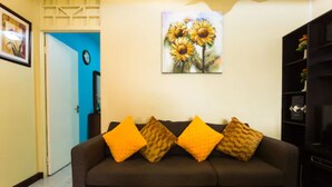 A living room with a dark brown sofa, three orange cushions, a painting of sunflowers, and a black bookshelf. - New Kingston Most Central Guest Apt (Kingston)