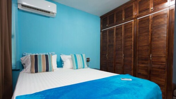 A bedroom with a bed, a blue blanket, striped pillows, a wooden wardrobe, and an air conditioning unit. - New Kingston Most Central Guest Apt (Kingston)