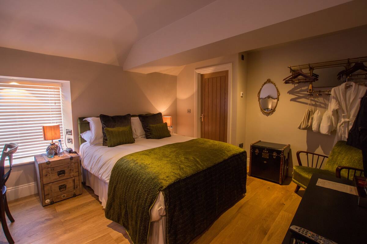 A bedroom with a bed, bedside table, lamp, dresser, and a coat rack with clothes.