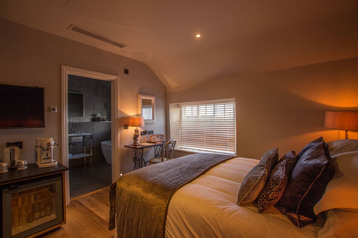 A bedroom with a bed, a desk with a chair, a window with blinds, and a kitchenette in the background.