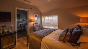 A bedroom with a bed, a desk with a chair, a window with blinds, and a kitchenette in the background. - The Peacock at Barlow (Dronfield)