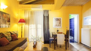 A living room with a sofa, a table with a vase, a lamp, and a dining area with chairs and a table. - Villa Kirki Apartments (Kefalonia)