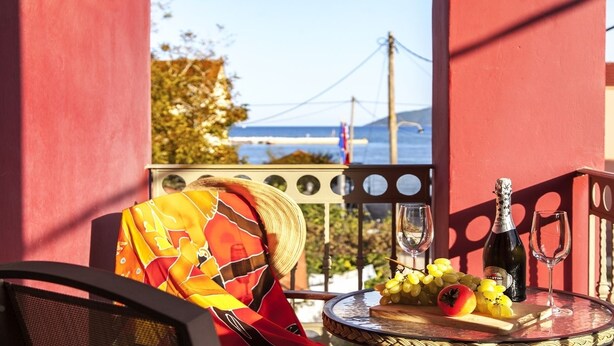 A balcony with a table set for a meal, featuring a bottle of wine, a glass, and a plate with grapes and a tomato. - Villa Kirki Apartments (Kefalonia)