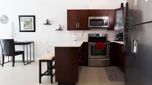 Una cucina moderna con mobili marrone scuro, elettrodomestici in acciaio inossidabile, un piano di lavoro bianco e una zona pranzo con una sedia e un tavolo neri.