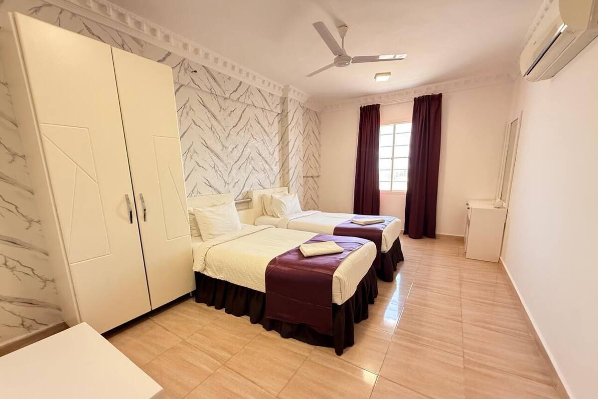 Habitación de hotel con dos camas, un armario, un ventilador de techo y una ventana con cortinas.
