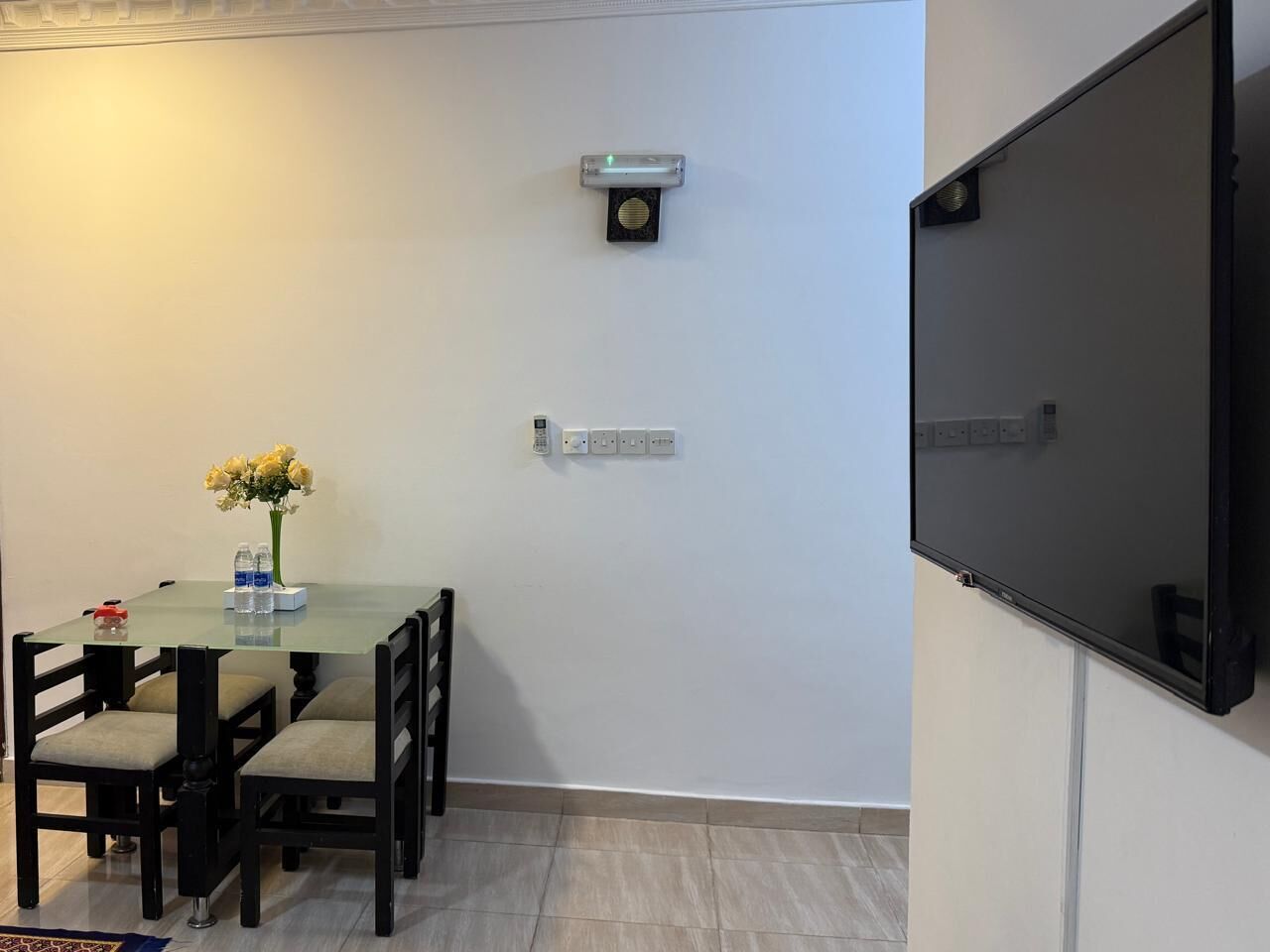 A small meeting room with a glass table, four chairs, a vase of flowers, a projector screen, and a wall-mounted light fixture.