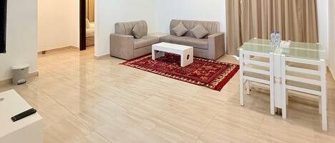 A spacious living room with a beige sofa, a white coffee table, and a glass dining table with chairs. There is a red patterned rug on the floor, a ceiling fan, and a painting on the wall.