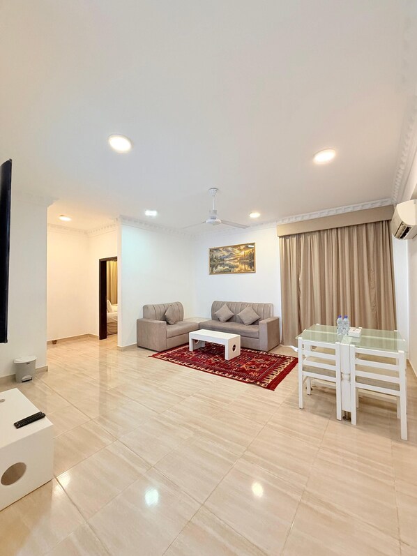 A spacious living room with a beige sofa, a white coffee table, and a glass dining table with chairs. There is a red patterned rug on the floor, a ceiling fan, and a painting on the wall. - Gateway Salalah (Salalah)