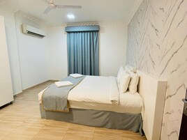 Habitación de hotel con cama, ventilador de techo, equipo de aire acondicionado, ventana con cortinas azules y pared con diseño de mármol.