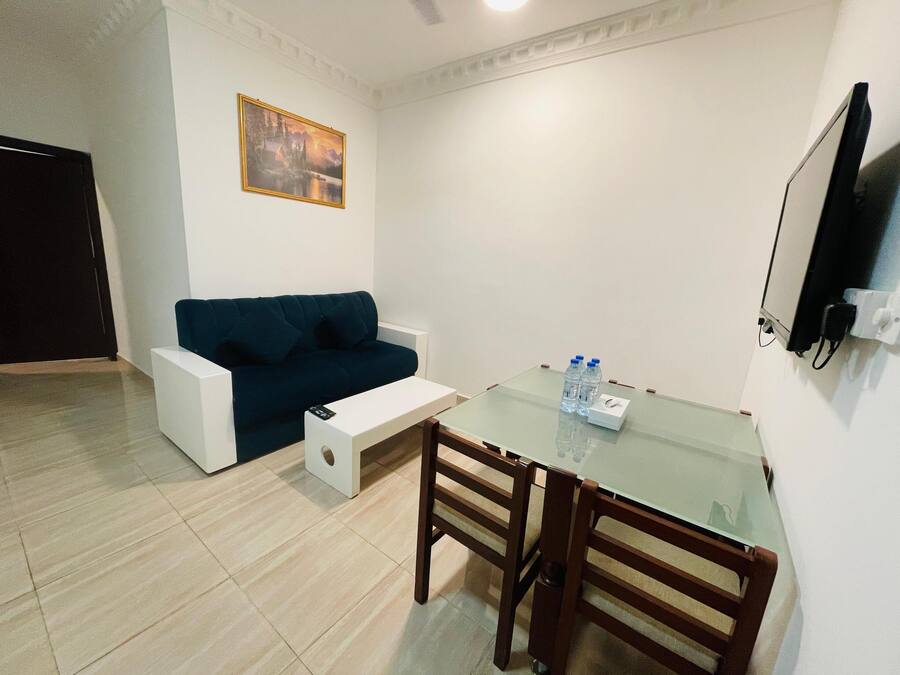 A small, modern living room with a dark blue sofa, a glass-top dining table with wooden chairs, and a flat-screen TV mounted on the wall.