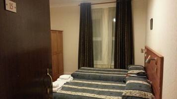 A hotel room with two beds, a wooden wardrobe, a window with curtains, and a door with a number sign.