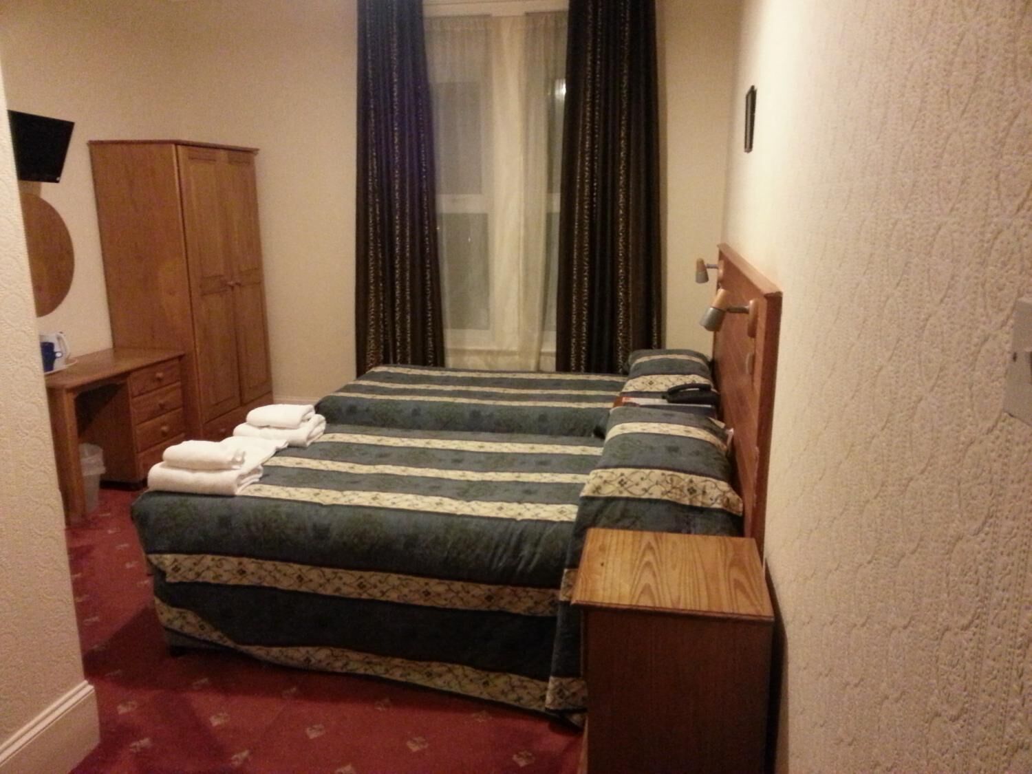 Una camera d'albergo modesta con due letti, un armadio in legno, una scrivania e una finestra con tende.