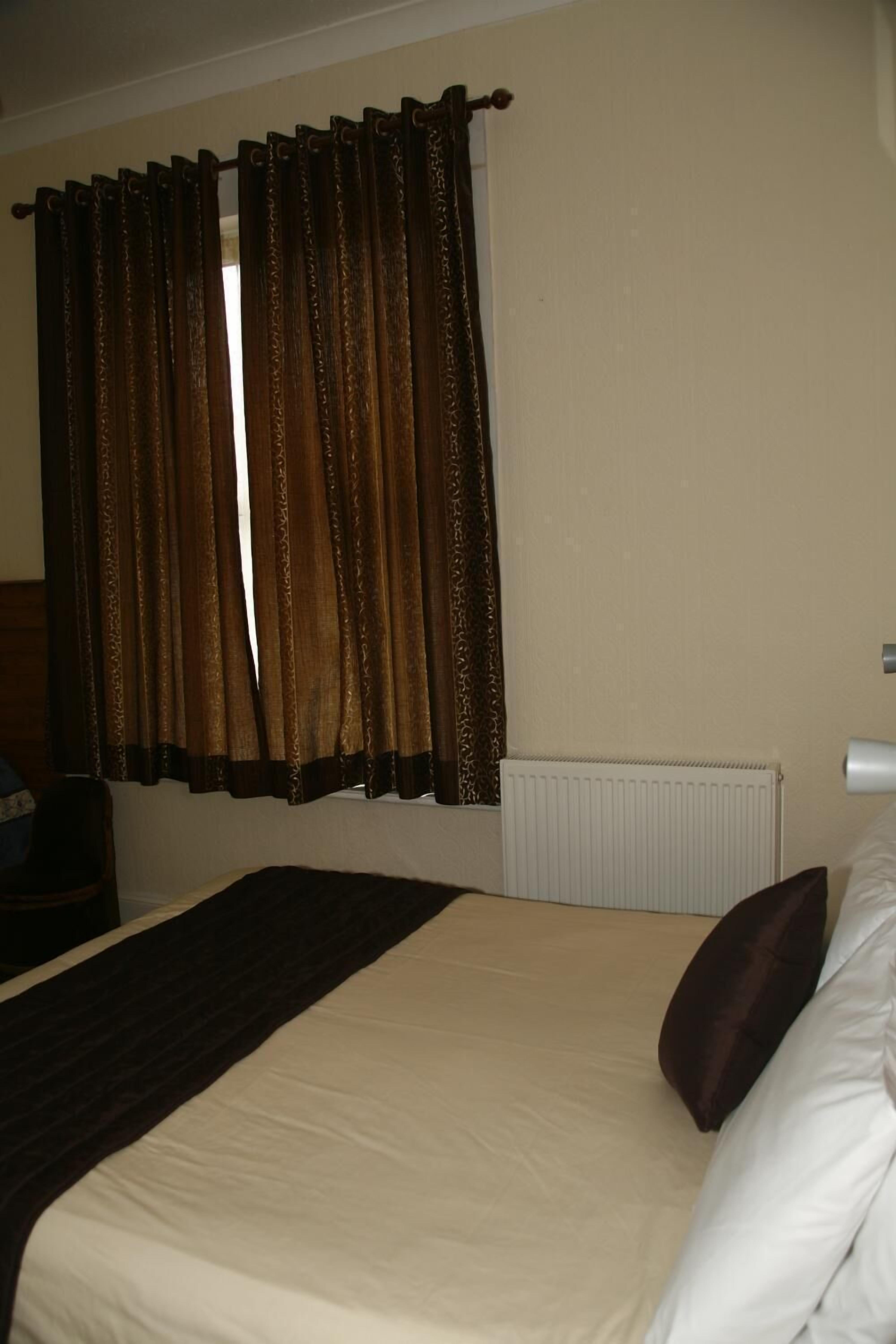 A hotel room with a bed, a radiator, a nightstand, and a window with curtains.