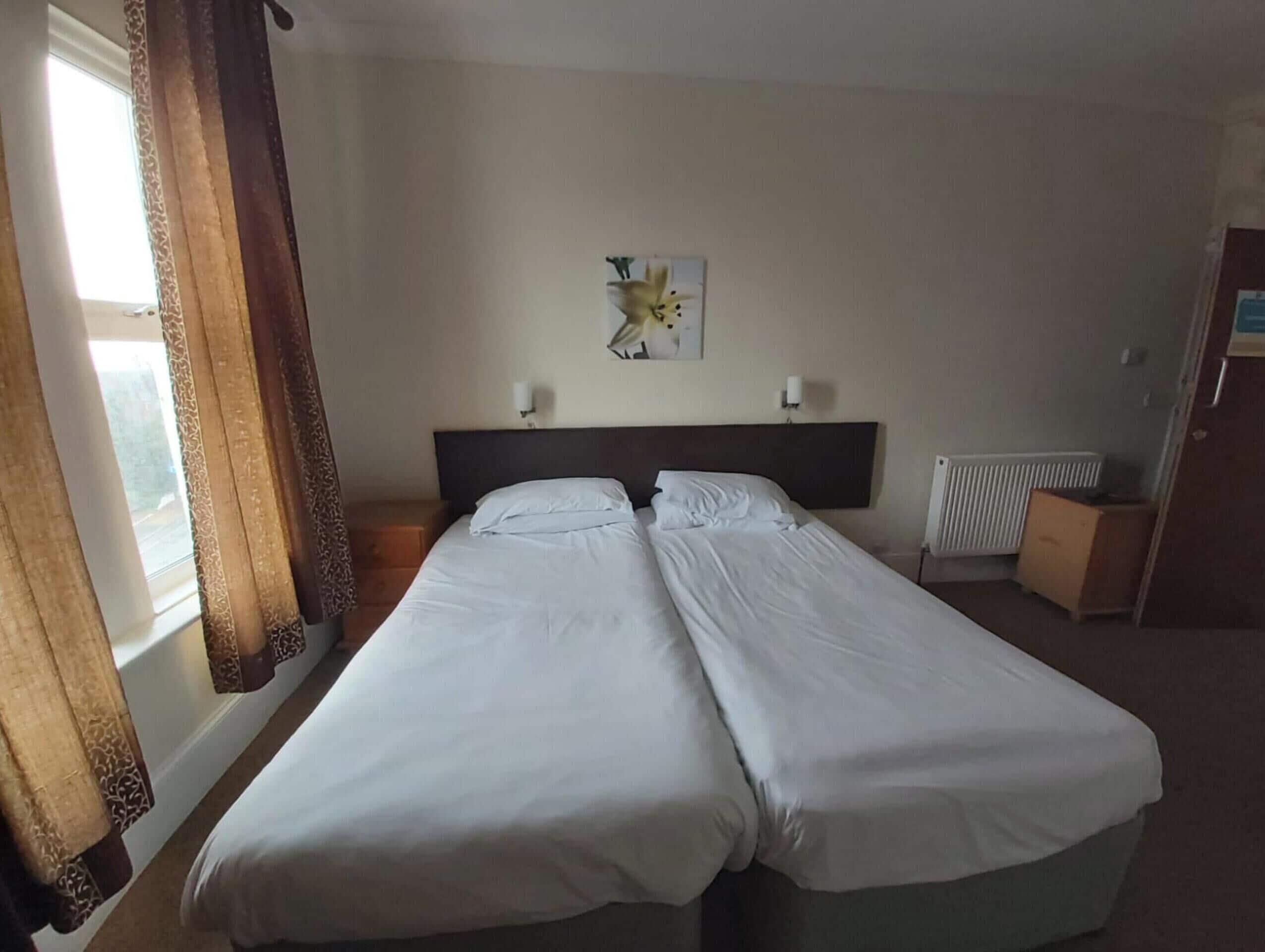 Una camera d'albergo con due letti singoli, una finestra con tende, un termosifone e una piccola credenza.