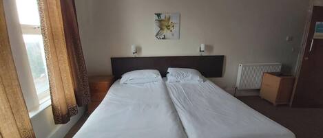 A hotel room with two single beds, a window with curtains, a radiator, and a small cabinet.