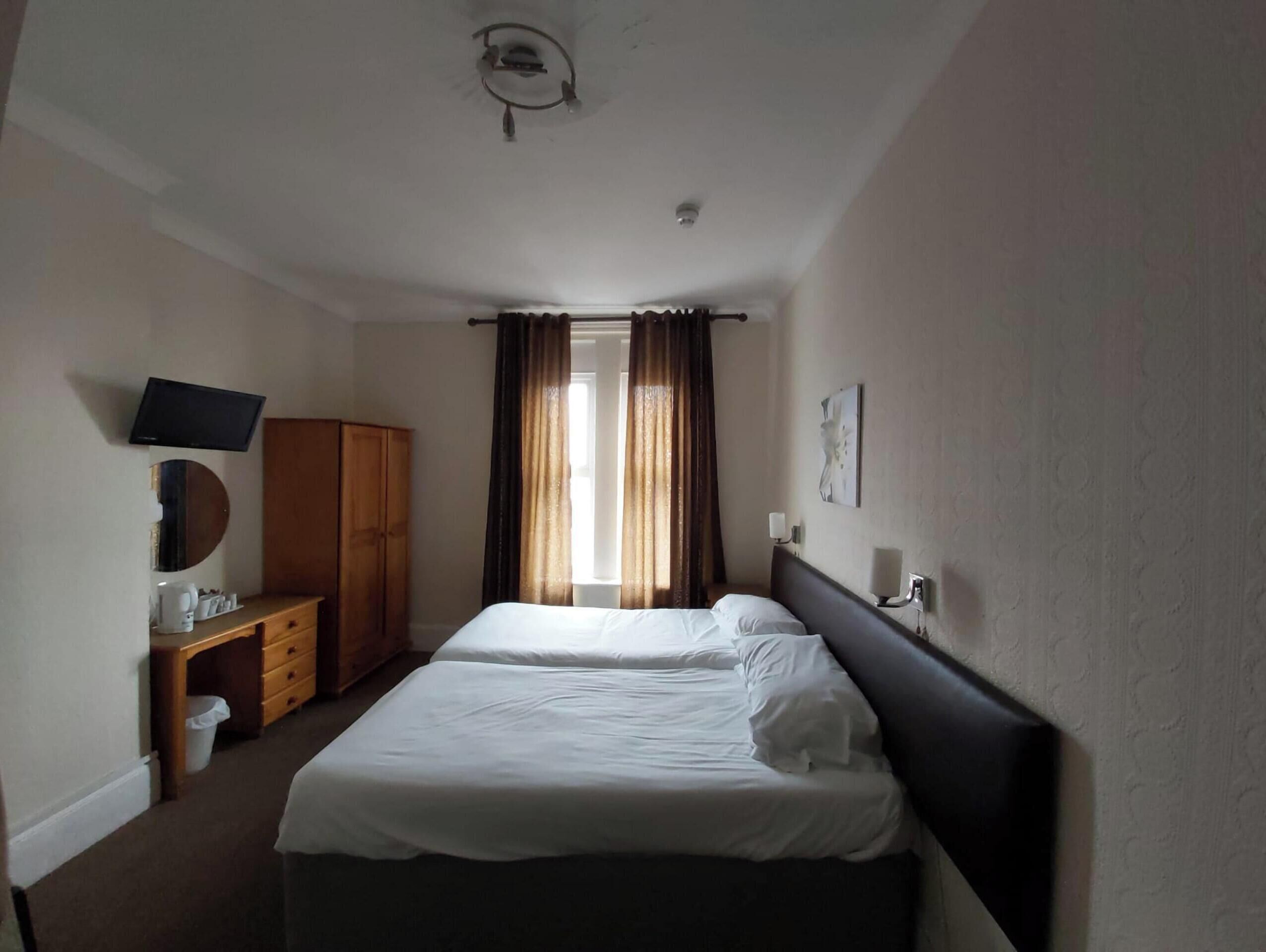 A hotel room with two single beds, a TV, a wooden wardrobe, and a desk with a chair.