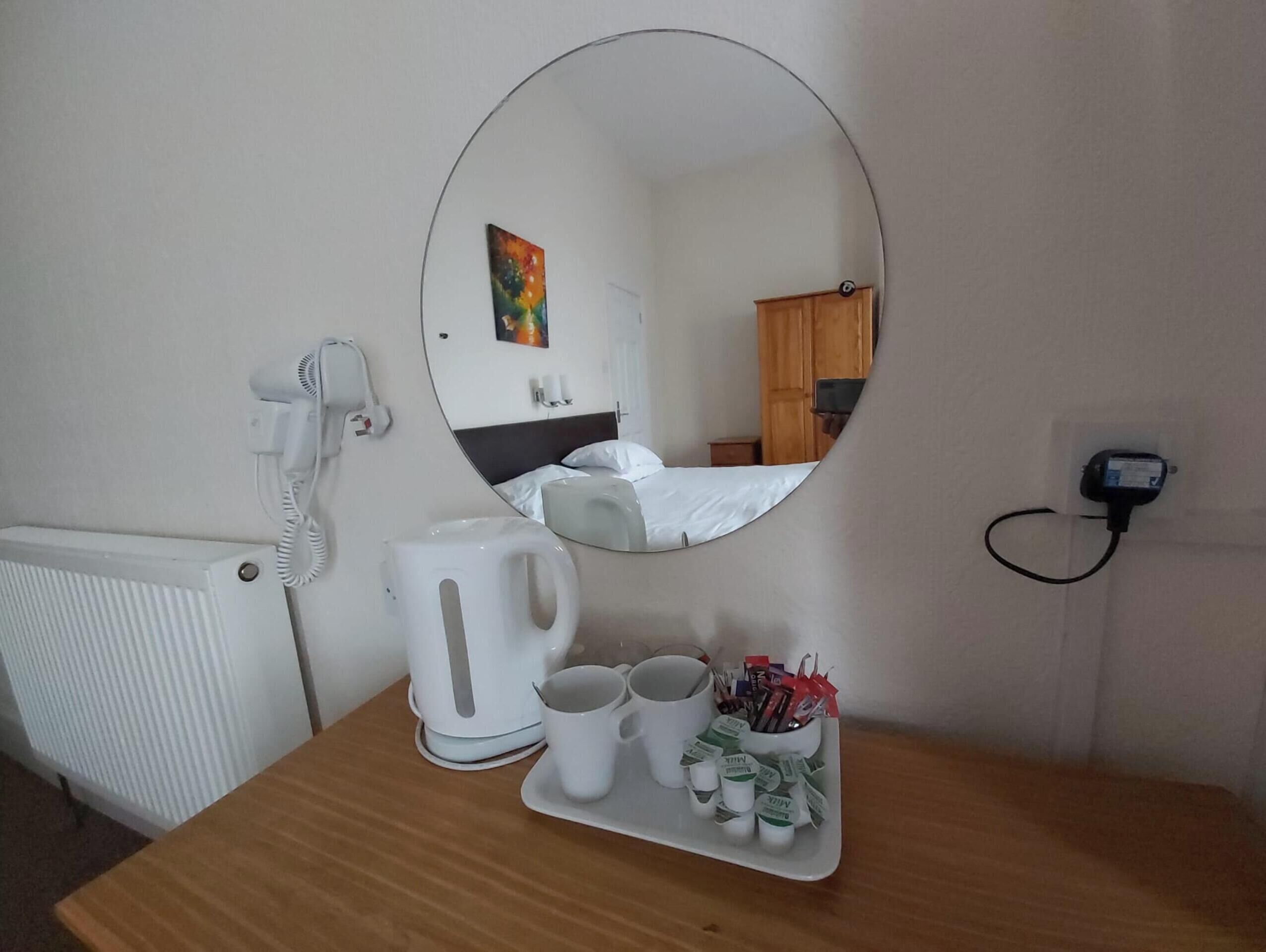 Habitación de hotel con un espejo redondo, un hervidor blanco, dos tazas y una bandeja con bolígrafos y blocs de notas.