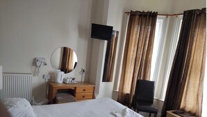A hotel room with a large bed, a wooden desk with a mirror, a chair, a TV mounted on the wall, and a window with curtains. - Channel View Hotel (Sandown)
