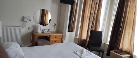 A hotel room with a large bed, a wooden desk with a mirror, a chair, a TV mounted on the wall, and a window with curtains.