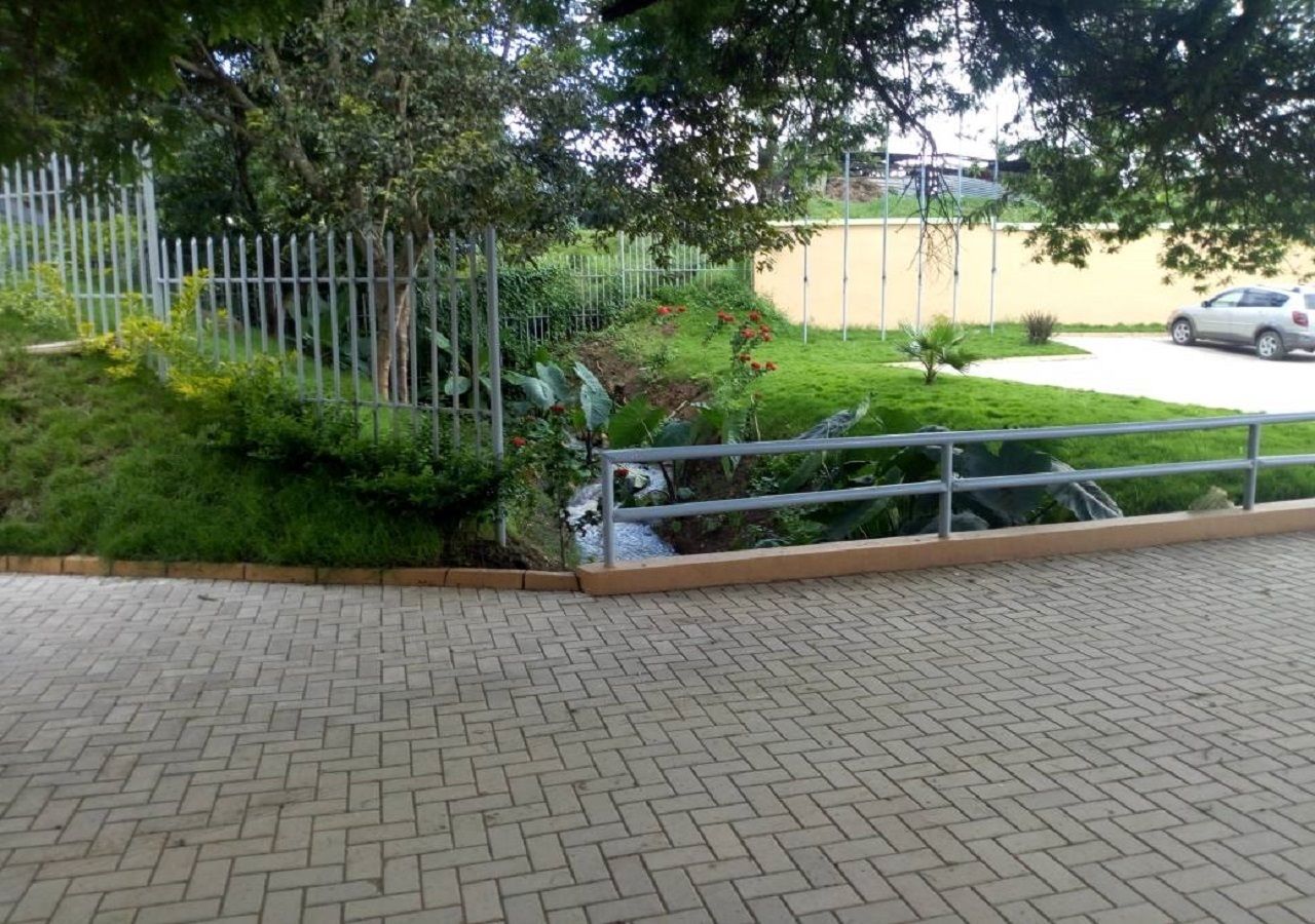 A paved walkway with a metal railing, surrounded by greenery and a fence.
