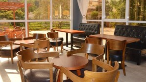Um café acolhedor com mesas e cadeiras de madeira, um sofá de couro preto e amplas janelas com vista para áreas verdes e edifícios.