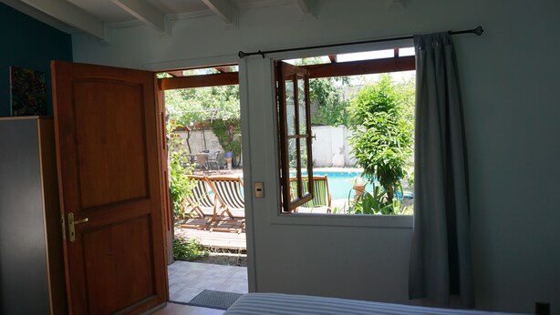 A bedroom with a wooden door, a window with curtains, a bed, and a view of an outdoor area with a pool and furniture. - Ventana Sur Hostel (Santiago)