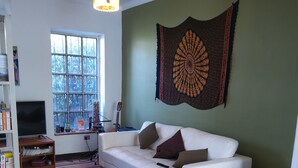 A living room with a white sofa, wooden chairs, a television, and a window with a view of trees. - Ventana Sur Hostel (Santiago)
