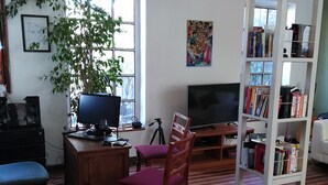 A room with a wooden floor, a desk with a computer, a chair, a bookshelf, and a large window with a plant. - Ventana Sur Hostel (Santiago)