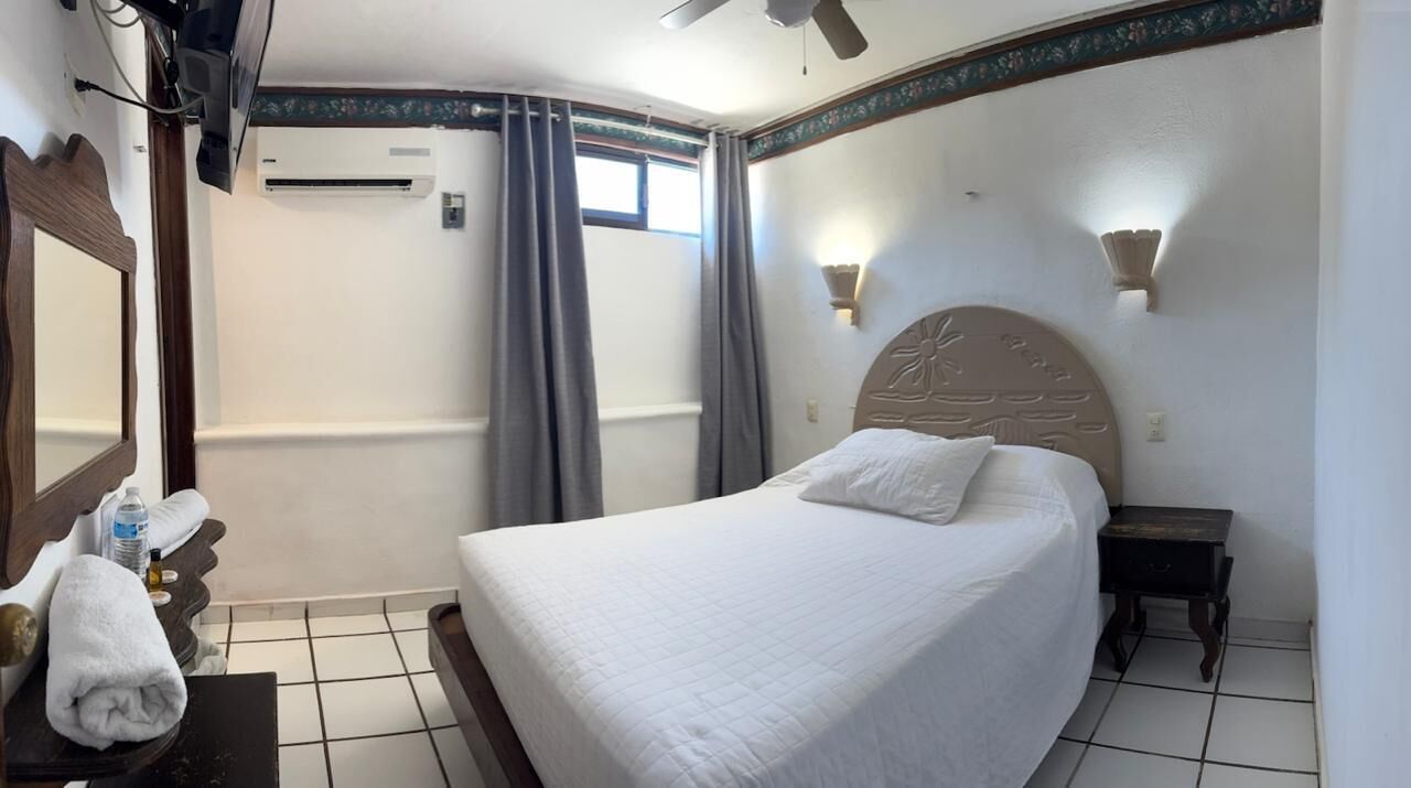 Habitación de hotel con cama, mesita de noche, espejo, ventilador de techo y ventana con cortinas.