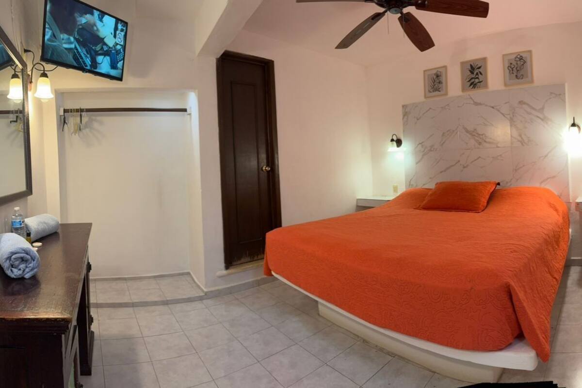A bedroom with a bed, a desk, a mirror, a ceiling fan, and wall-mounted lights.