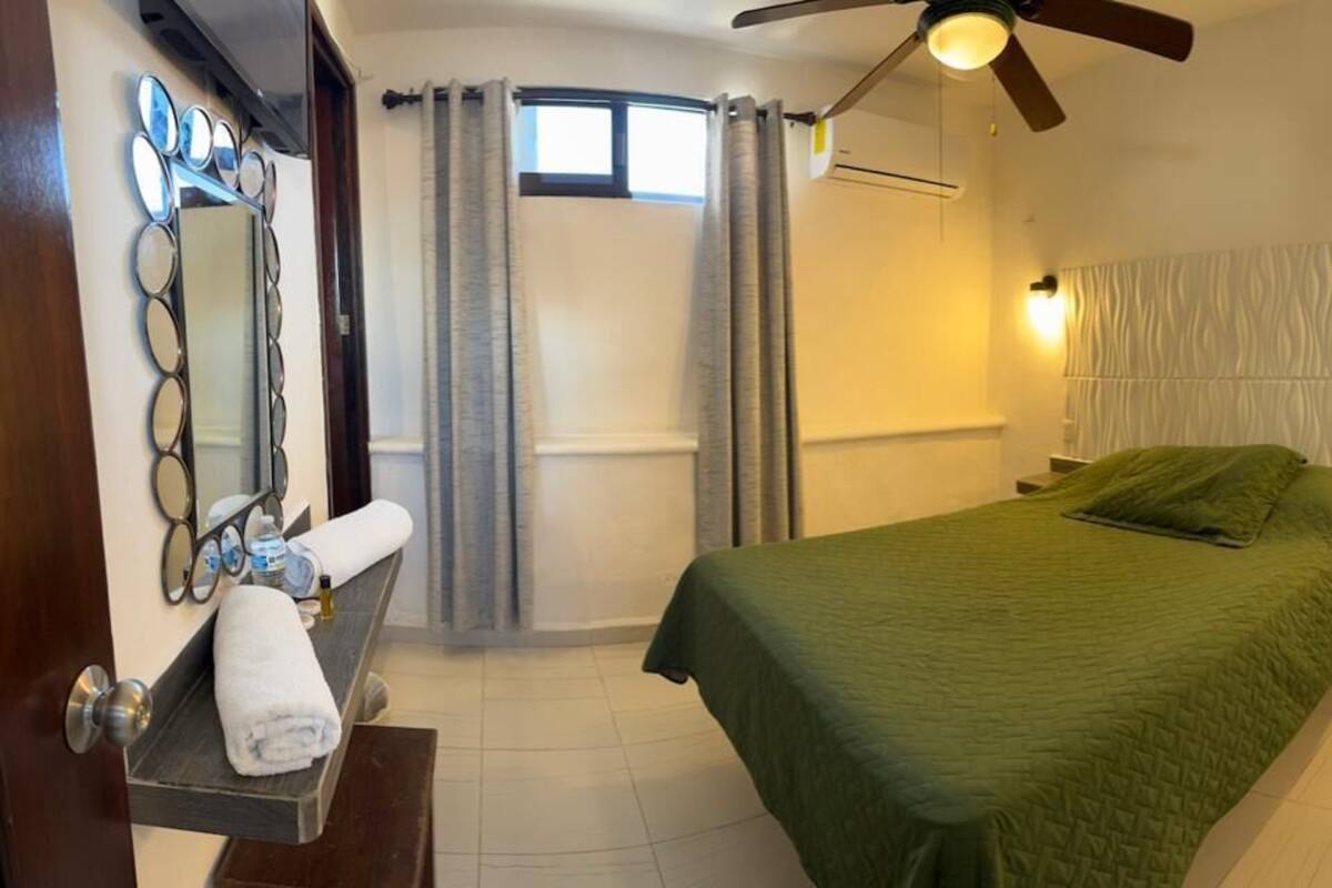 A bedroom with a bed, a ceiling fan, a nightstand, a mirror, and a window with curtains.