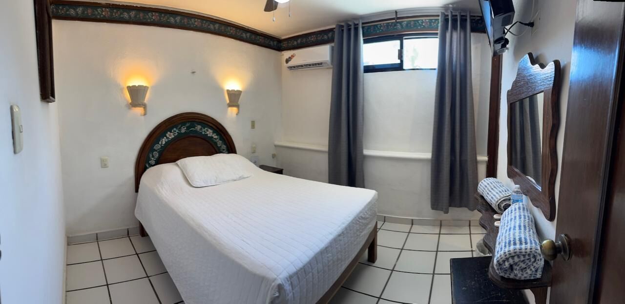Una camera d'albergo con un letto, due luci a parete, una finestra con tende, uno specchio e un portascopevoli.
