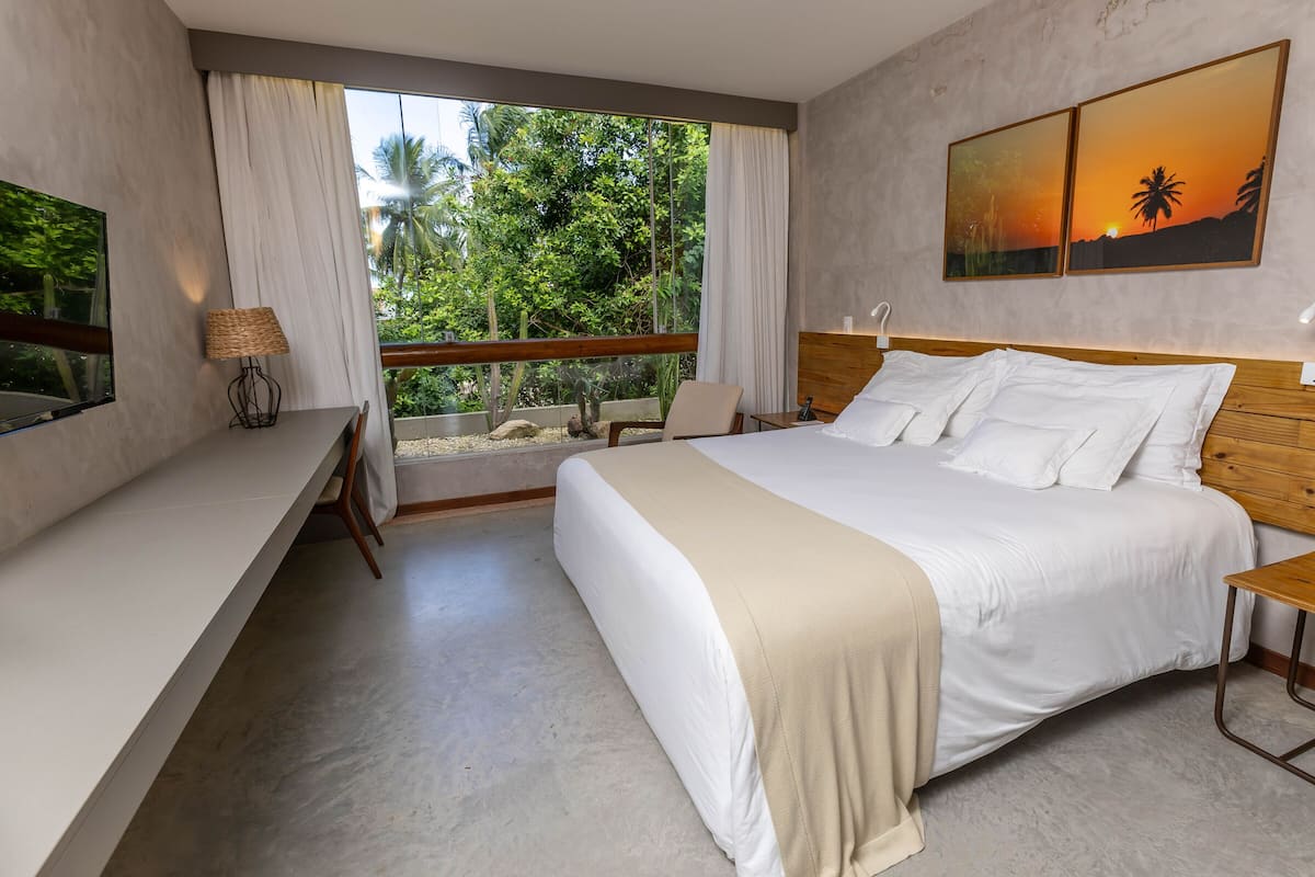 A modern hotel room with a large bed, a wooden headboard, a desk, and a view of lush greenery through a large window.