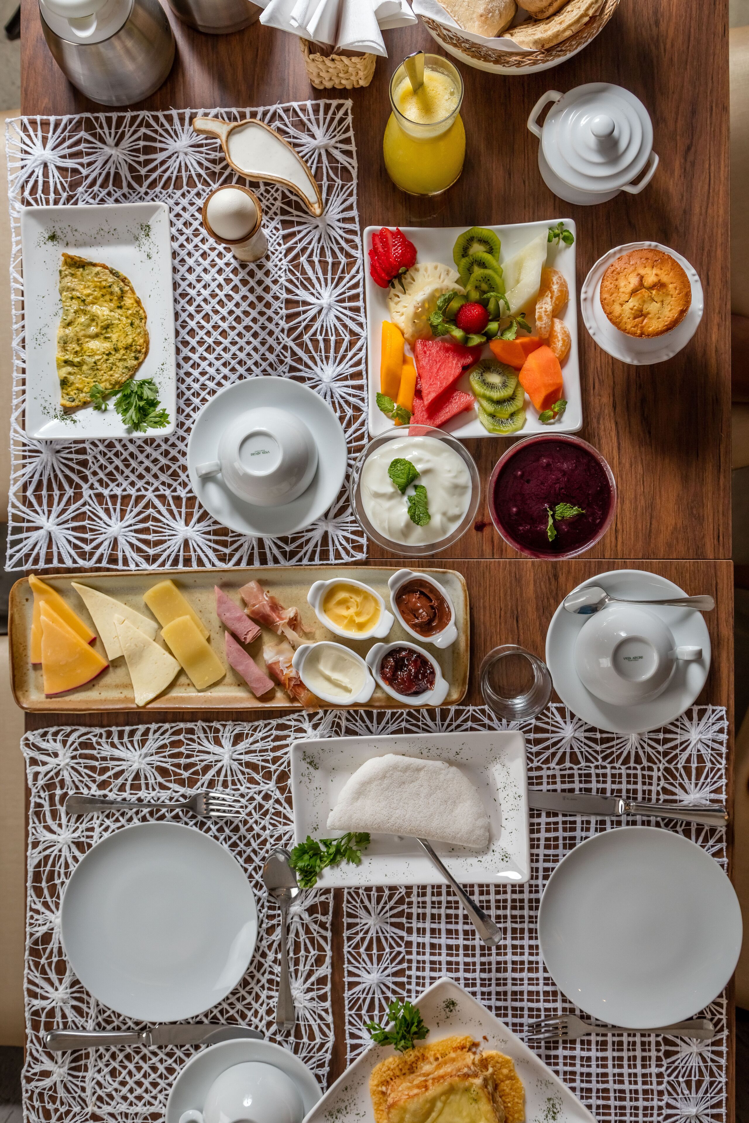 Un desayuno con diversas opciones de comida, como una tortilla, fruta, queso y pastelería, dispuestas sobre una mesa de madera con mantel blanco estampado.