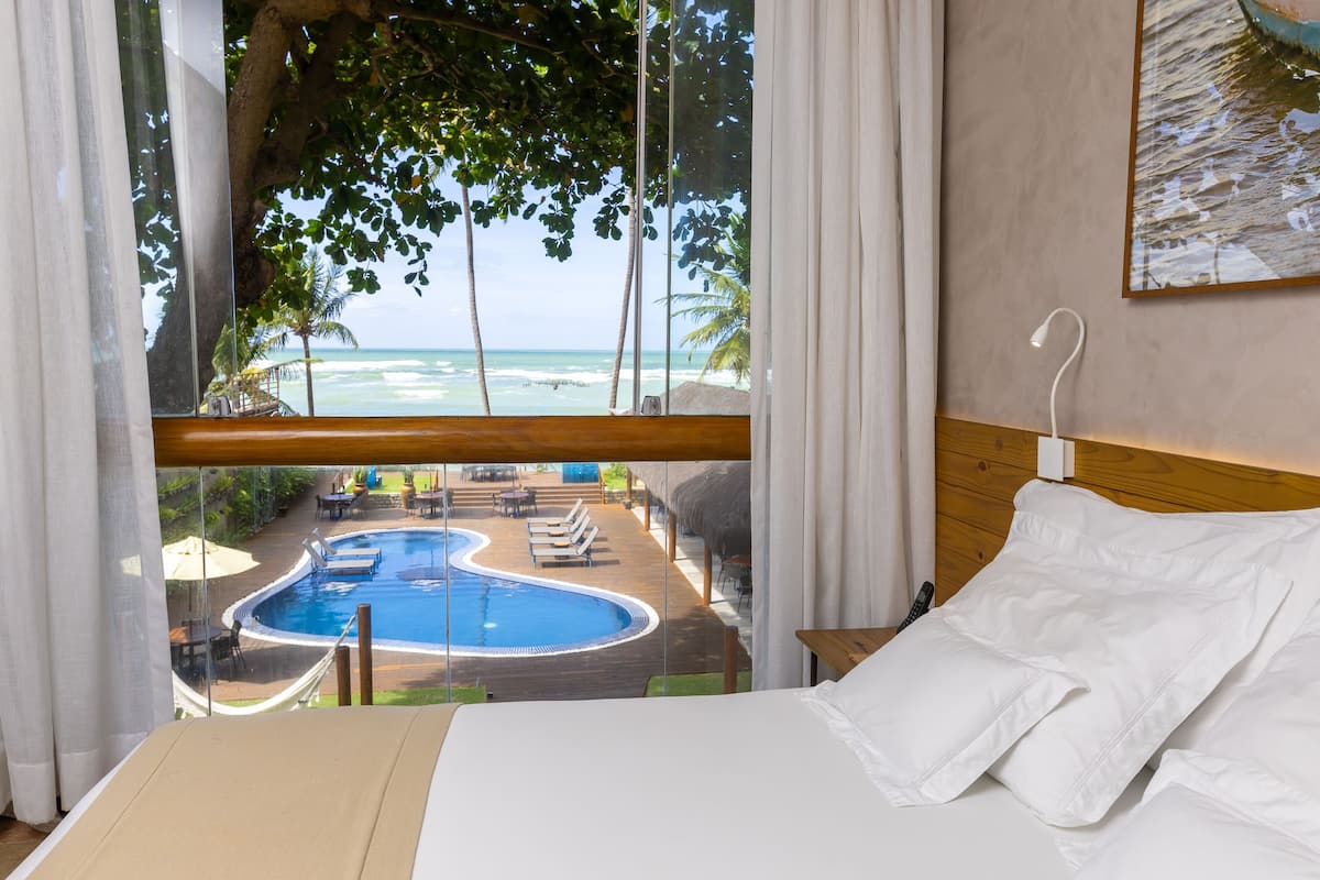 A hotel room with a bed, a view of a swimming pool, and a beach.