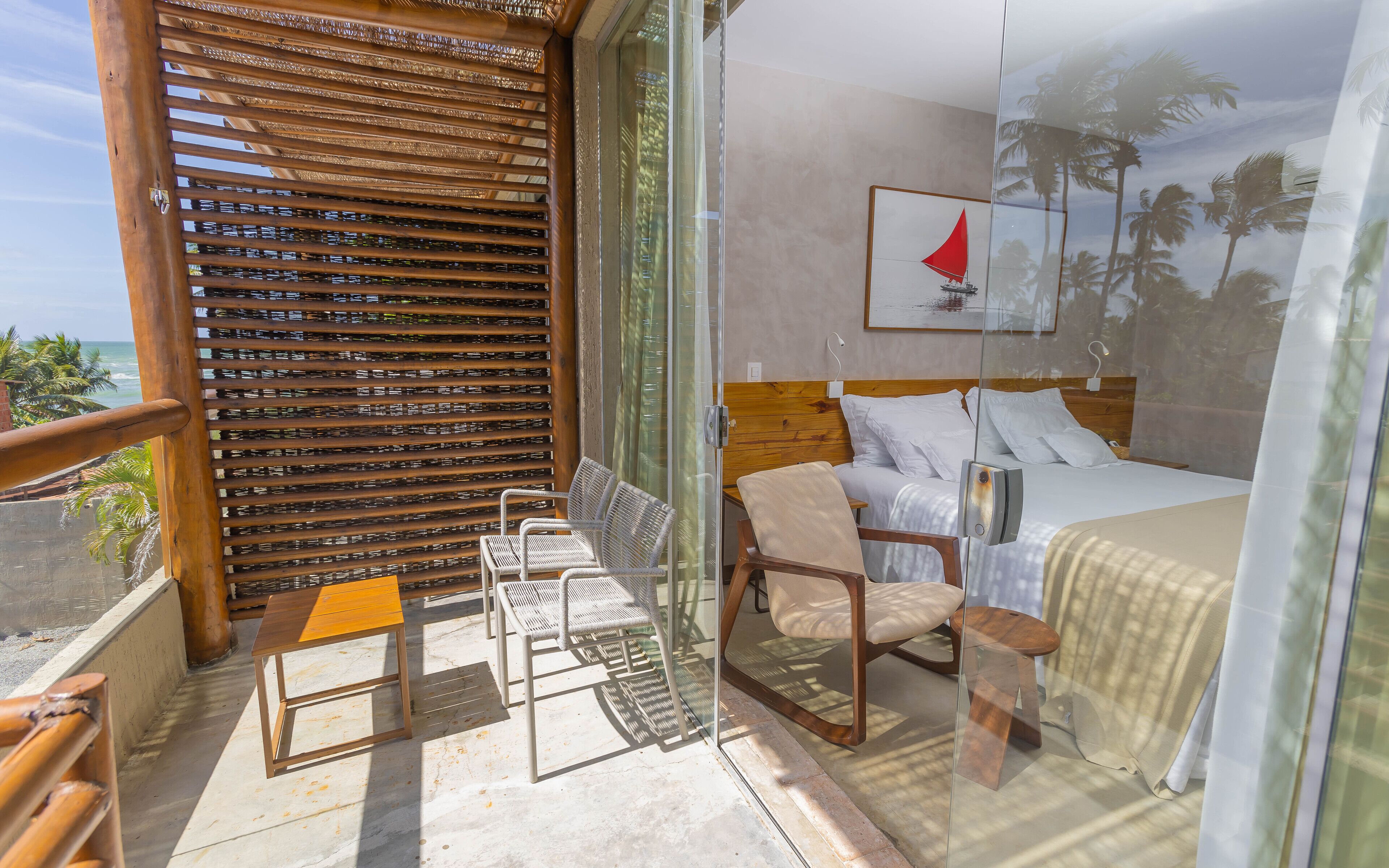 Un balcón con una celosía de madera, una cama con ropa blanca y una mesita con una silla.