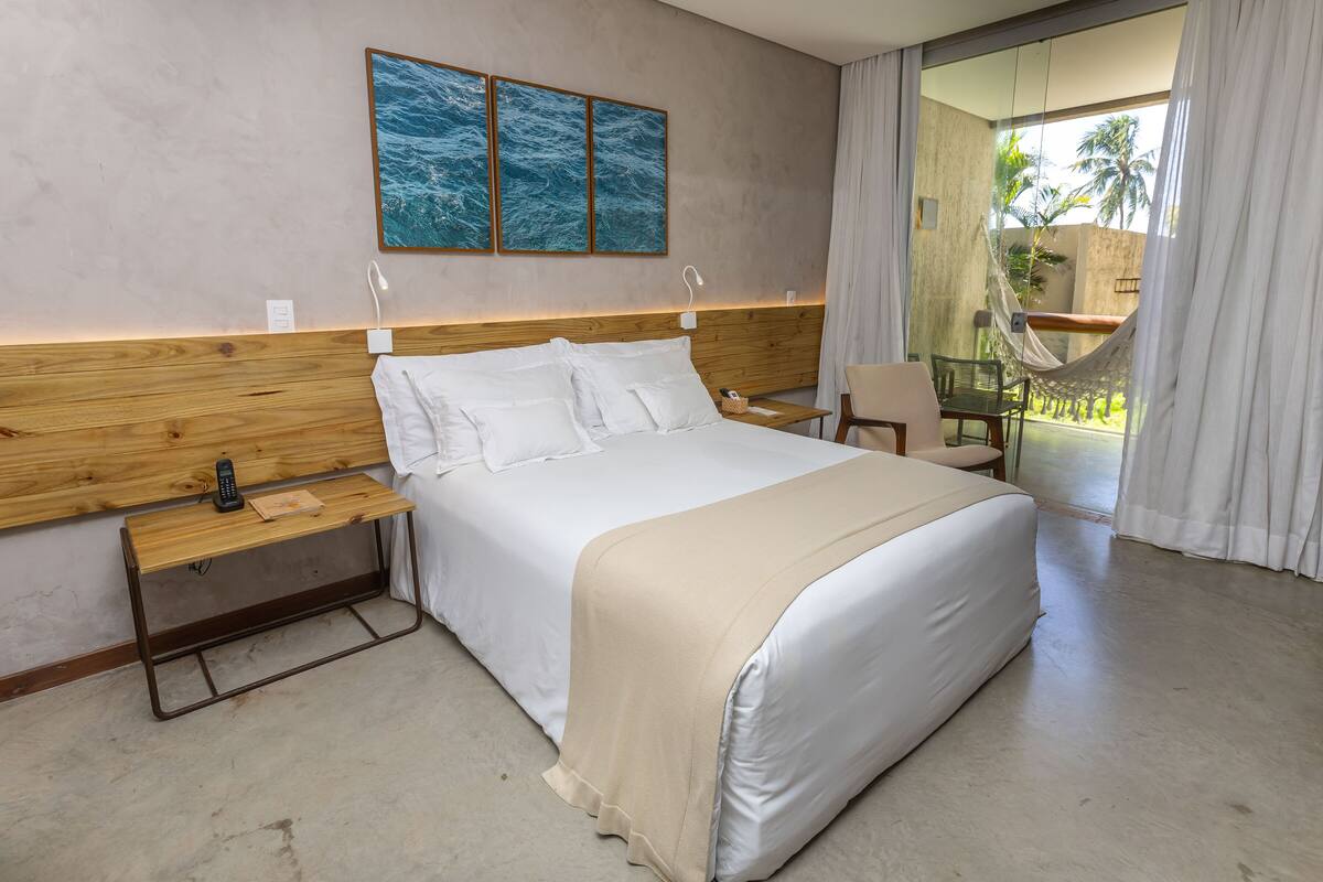 A modern hotel room with a large bed, wooden headboard, and a view of a balcony with a hammock.