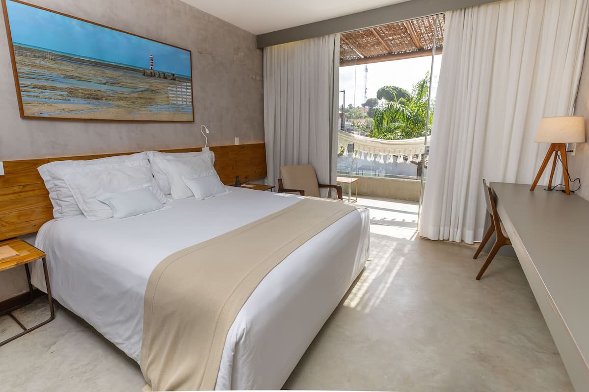 A hotel room with a large bed, a desk, a chair, and a balcony with a view of the beach and a lighthouse.