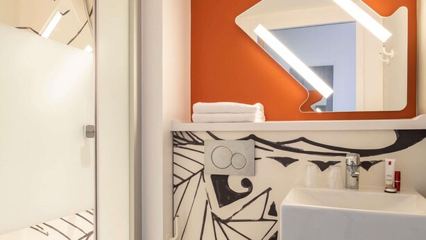 A modern bathroom with a white sink, a mirror with an orange accent wall, and a shower with a handheld showerhead. - ibis budget Strasbourg Centre Gare (Strasbourg)