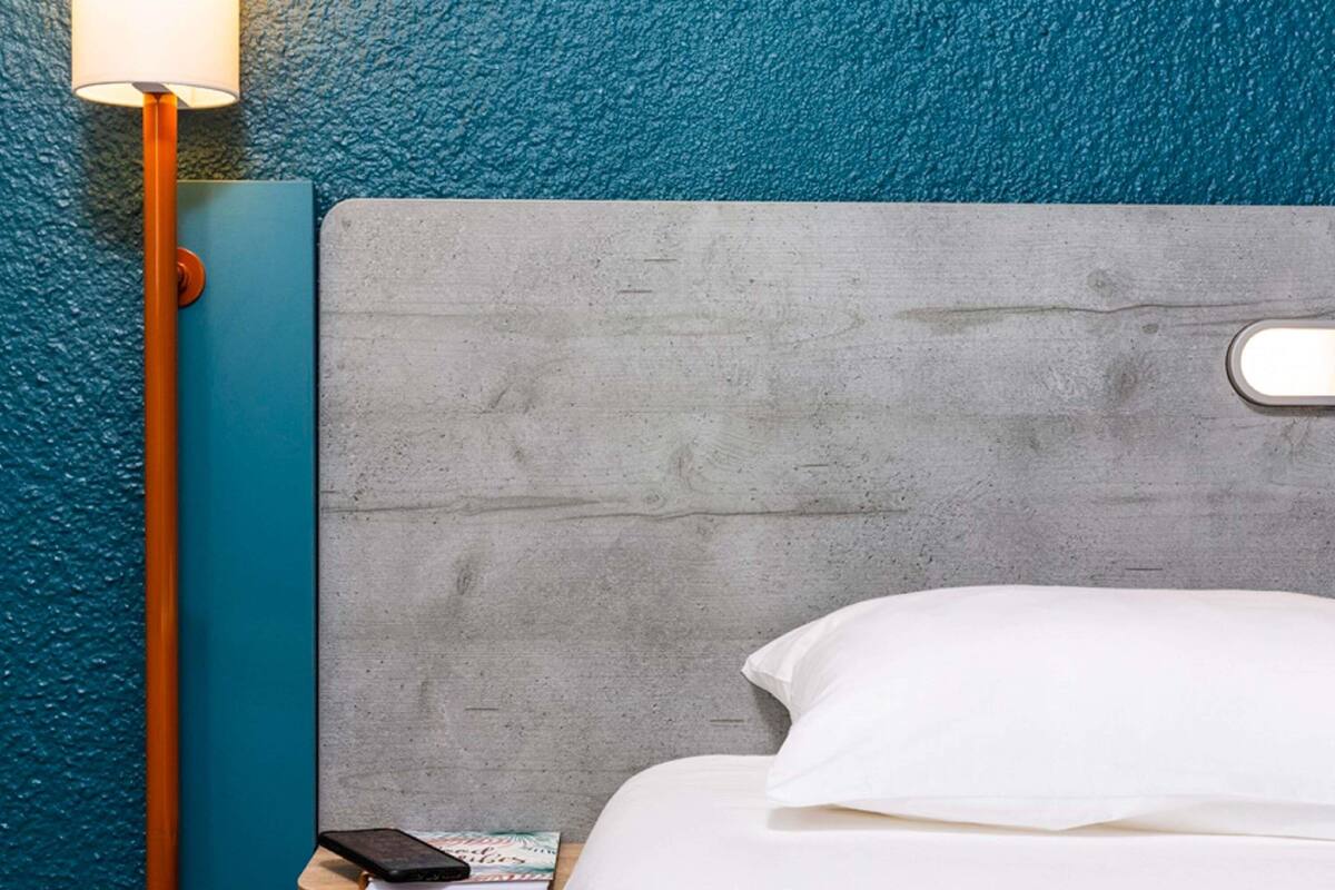 A hotel room with a concrete headboard, a bedside table with a lamp, a phone, and a book.