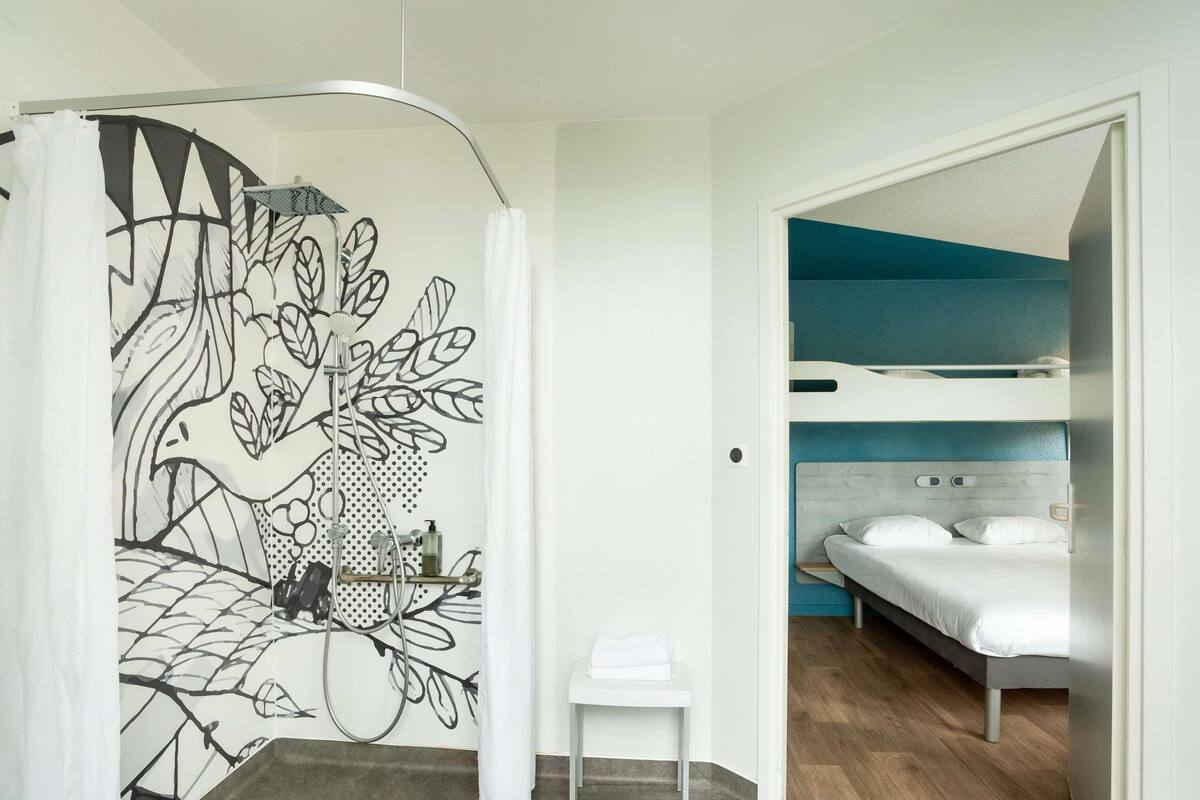 A room with a bunk bed, a shower with a leaf-patterned curtain, a small white table, and a grey carpet.