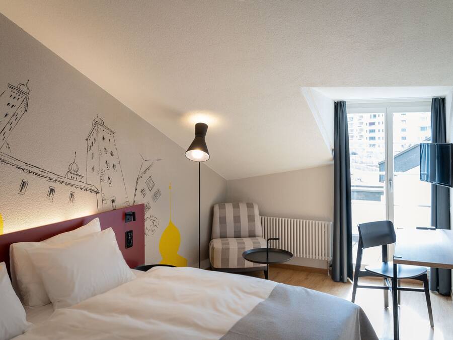 A hotel room with a bed, a chair, a small table, a lamp, and a wall with a cityscape mural.