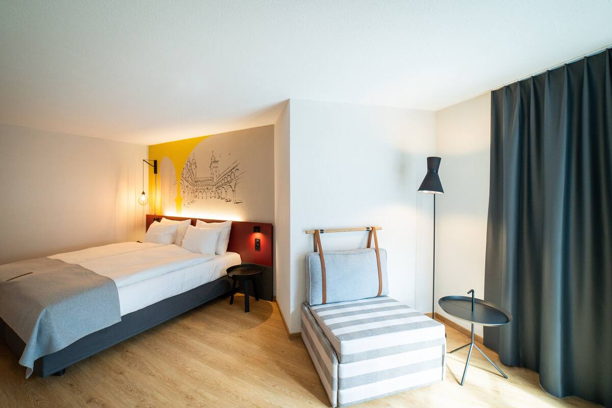 Una habitación de hotel con una cama grande, una cama más pequeña, una mesita de noche, una lámpara y una obra de arte en la pared.