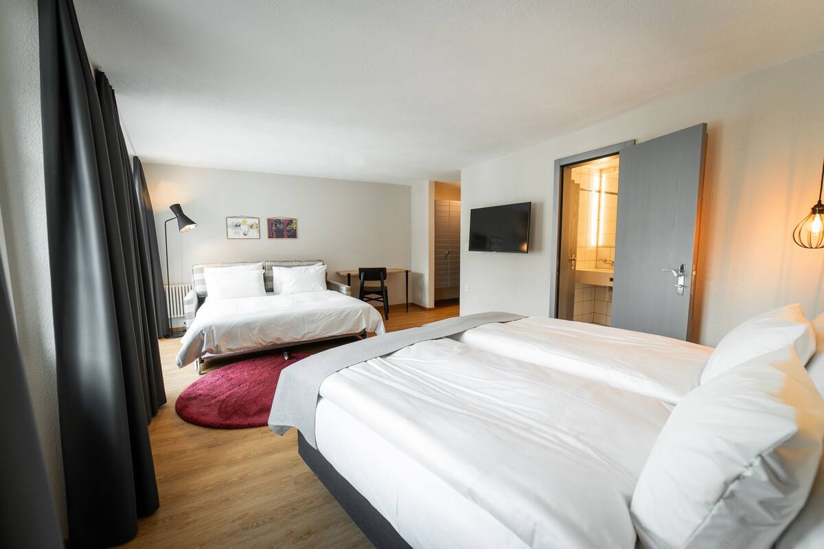 Habitación de hotel con dos camas, televisión, escritorio y baño visible a través de una puerta abierta.