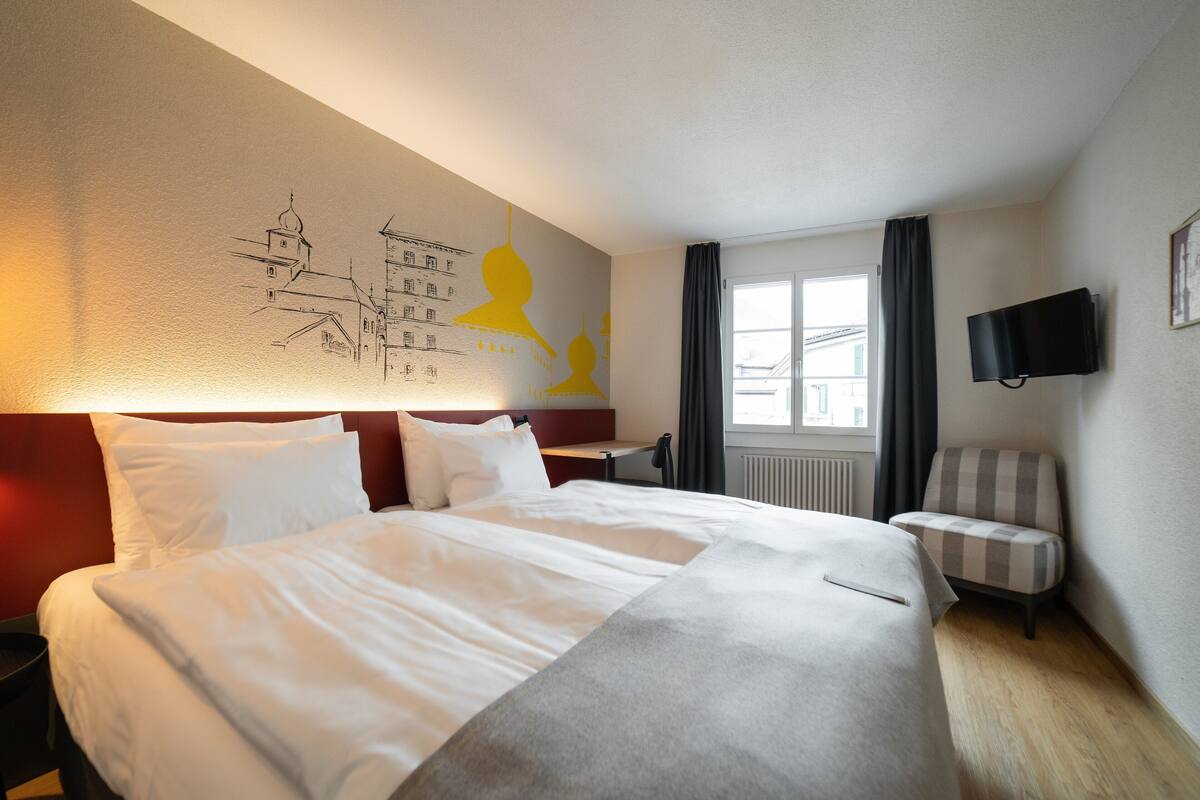 Habitación de hotel con una cama grande, un escritorio, una silla, un televisor y una ventana con cortinas.