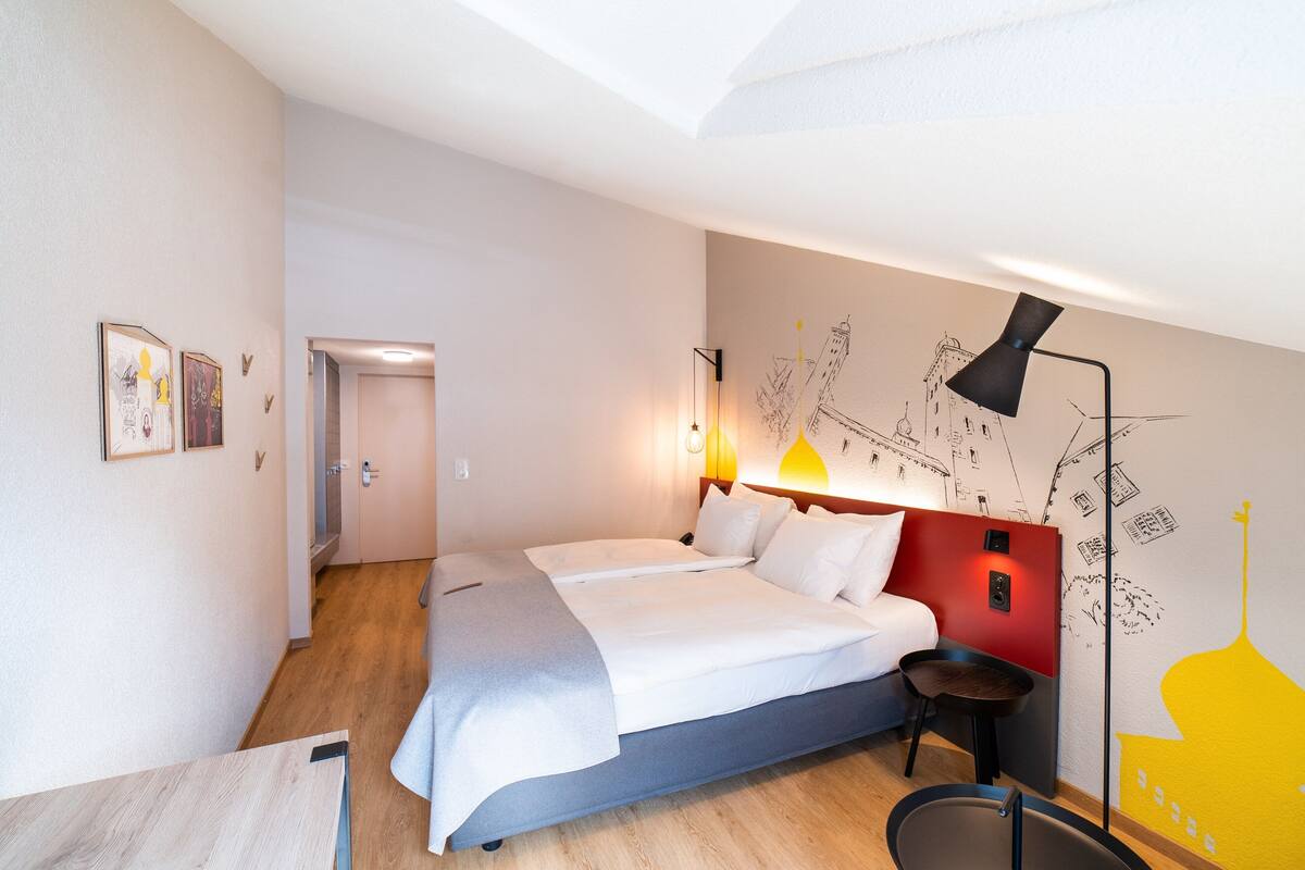 Una habitación de hotel moderna con una cama grande, un escritorio y una silla. Hay una puerta que conduce a otra habitación.